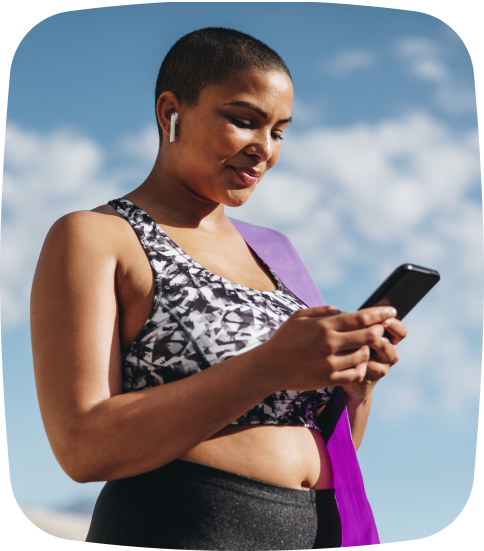 A woman in yoga attire uses her phone outdoors, smiling as she books a class through her studio's mobile app—highlighting the ease and convenience TeamUp provides for modern yoga clients. This seamless mobile experience showcases the value of using a user-friendly, mobile-optimized Yoga Class Booking Software that keeps customers engaged and classes full.